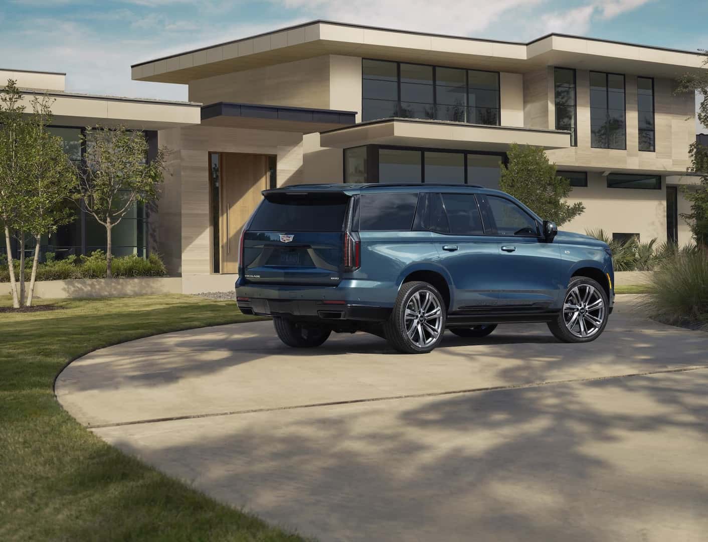 Rear ¾ view of the 2025 Cadillac Escalade Sport Platinum in Deep Sea Metallic with a house and greenery in the background.