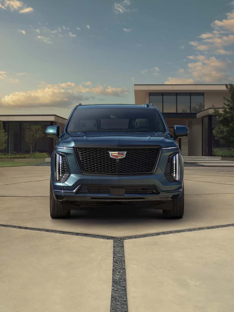 Head-on view of the 2025 Cadillac Escalade Sport Platinum in Deep Sea Metallic with a house and greenery in the background.