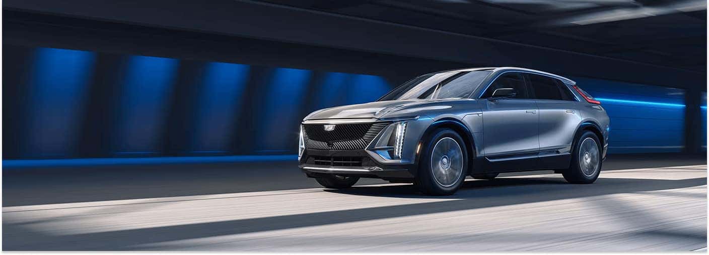 Silver 2026 Cadillac LYRIQ electric SUV driving through a tunnel with illuminated front grille and vertical LED lights.