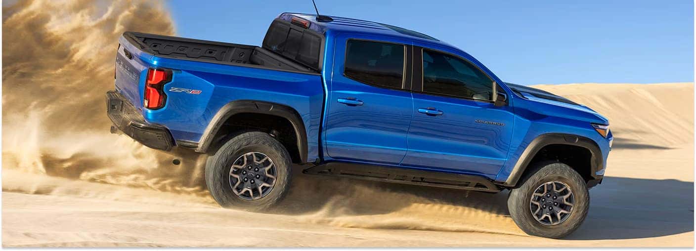 Blue 2026 Chevrolet Colorado ZR2 pickup truck driving through desert sand dunes.