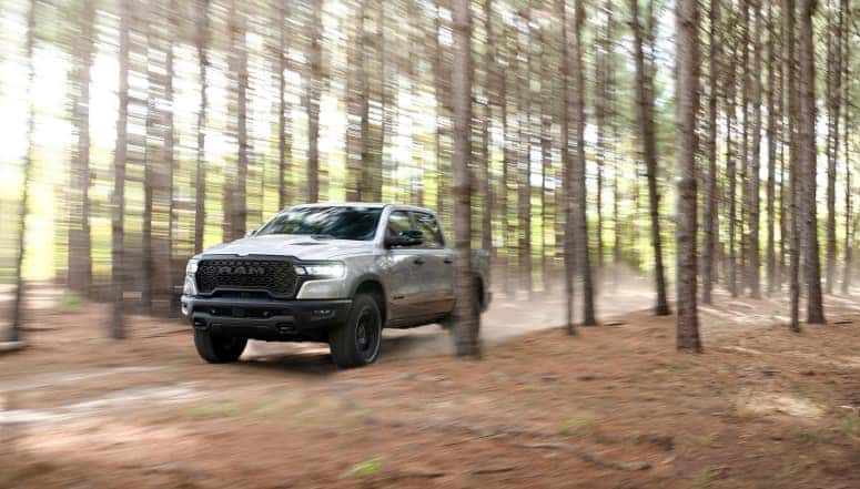 Ram 1500 (DT) Rebel in Silver Zynith driving on dirt road through a forest of trees.