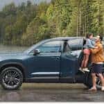 Side profile of a blue 2026 Toyota Grand Highlander parked by a lake with rear door open and trailer attached.