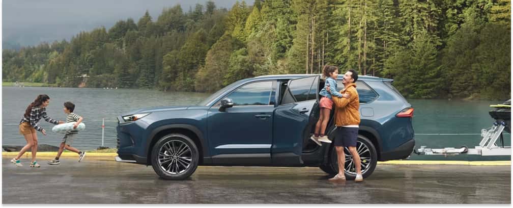 Side profile of a blue 2026 Toyota Grand Highlander parked by a lake with rear door open and trailer attached.