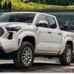 White 2026 Toyota Tacoma pickup truck towing a boat trailer at a lakeside boat launch.