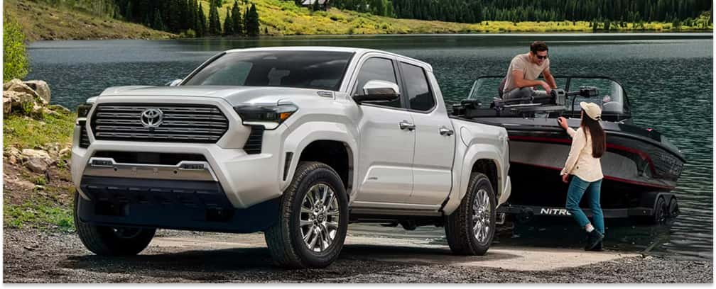 White 2026 Toyota Tacoma pickup truck towing a boat trailer at a lakeside boat launch.
