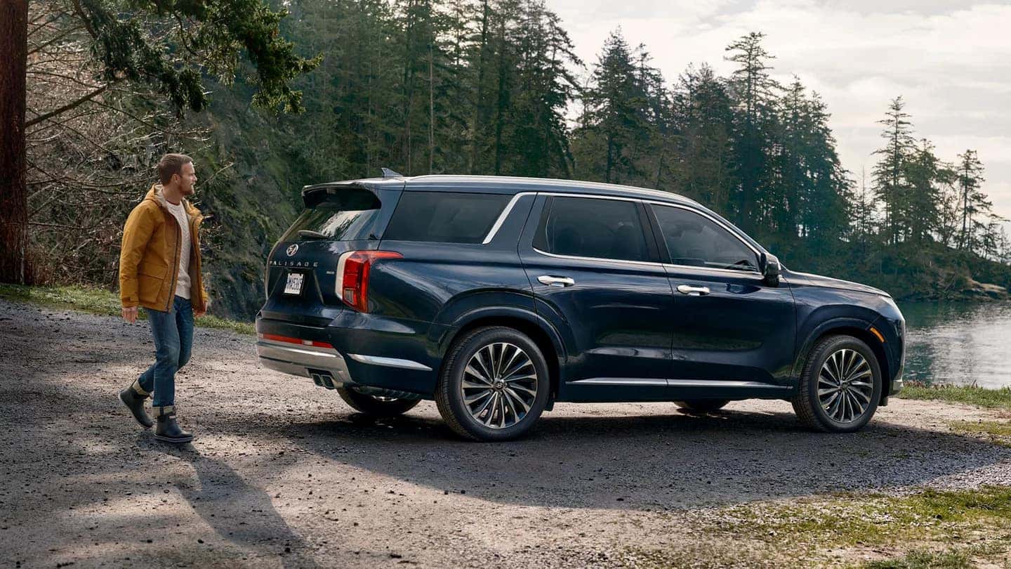 A blue Hyundai Palisade parked at a lake by a forest with a man walking behind it