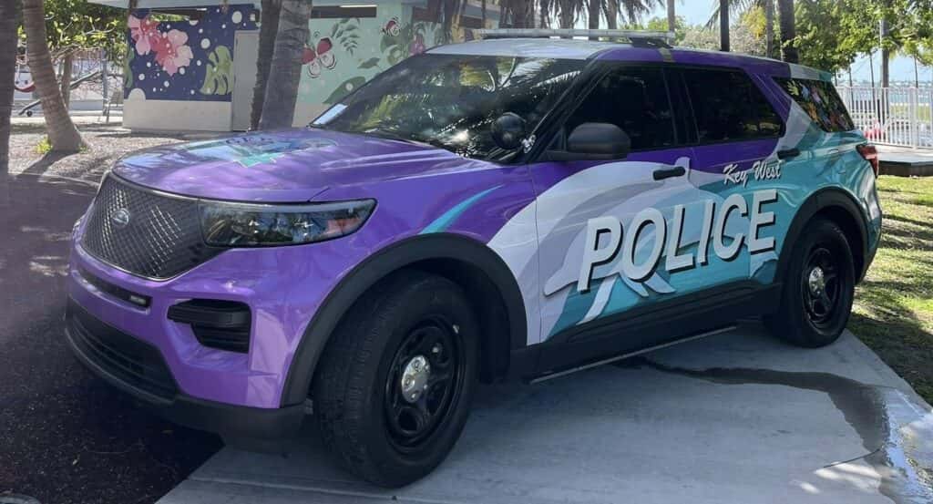 A brightly colored Key West Police SUV is parked outdoors near palm trees and a mural-covered building. The vehicle features a custom wrap in shades of purple, blue, and white with "POLICE" and "Key West" prominently displayed on the side.
