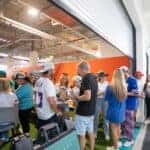 A crowd of guests socialize inside a covered lounge area during an exclusive tailgate event hosted by Land Rover North Dade, South Dade, and West Broward for the Miami Dolphins game. People wearing team jerseys and casual outfits enjoy food and drinks while gathered around tables and standing in small groups. The space features bright lighting, sports décor, and an energetic game-day atmosphere.