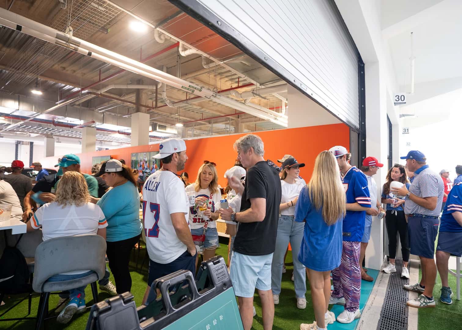 A crowd of guests socialize inside a covered lounge area during an exclusive tailgate event hosted by Land Rover North Dade, South Dade, and West Broward for the Miami Dolphins game. People wearing team jerseys and casual outfits enjoy food and drinks while gathered around tables and standing in small groups. The space features bright lighting, sports décor, and an energetic game-day atmosphere.