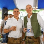 Two men wearing traditional lederhosen stand together at an Oktoberfest-themed charity event, smiling and holding drinks inside a festive tent with blue and white decorations. One wears a white polo shirt, and the other wears a white shirt with a green vest. Other attendees and musical equipment are visible in the background.