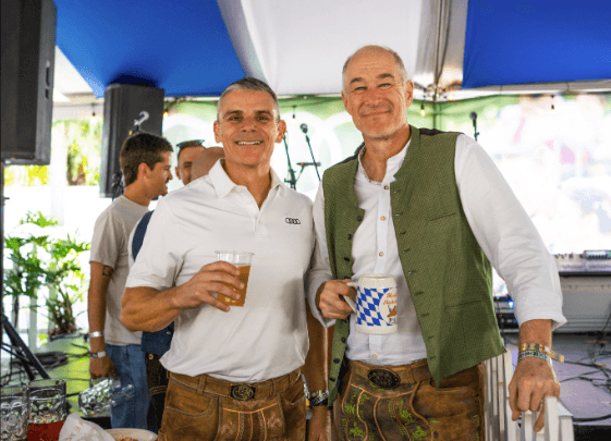 Two men wearing traditional lederhosen stand together at an Oktoberfest-themed charity event, smiling and holding drinks inside a festive tent with blue and white decorations. One wears a white polo shirt, and the other wears a white shirt with a green vest. Other attendees and musical equipment are visible in the background.
