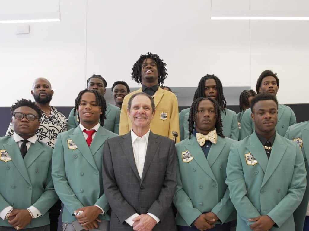 Warren Henry Zinn stands alongside the 2026 Tournament of Champions student-athletes during an awards ceremony, as the players pose together wearing commemorative sport coats recognizing their achievements on and off the football field.